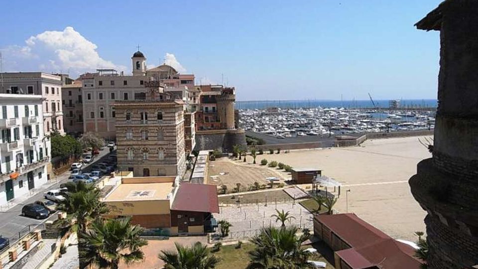 Veduta panoramica di un porto turistico sul mare con numerose barche ormeggiate, affiancato da una grande piazza aperta e da edifici storici lungo il lungomare. In primo piano si vedono palme, piccoli edifici con tettoie e una strada costeggiata da palazzi.