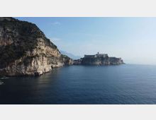 Veduta costiera con alte scogliere rocciose a picco sul mare calmo; in lontananza si vede un promontorio con una fortezza o complesso di edifici storici affacciati sull’acqua. Cielo sereno e costa frastagliata che prosegue verso l’orizzonte.