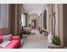 Corridoio interno di un edificio storico con soffitto a volte, colonne e pavimento piastrellato chiaro. Lungo le pareti ci sono finestre alte, lanterne, quadri e decorazioni con vasi, piante, tappeti e una zona divano rossa; in fondo si vede una porta aperta.