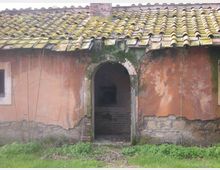Facciata di un edificio rurale con intonaco scrostato e tetto in coppi, con un arco centrale che conduce a un ingresso buio con gradini interni. Alla base si vede terreno erboso con vegetazione bassa e tracce di umidità e muschio attorno all’arco.