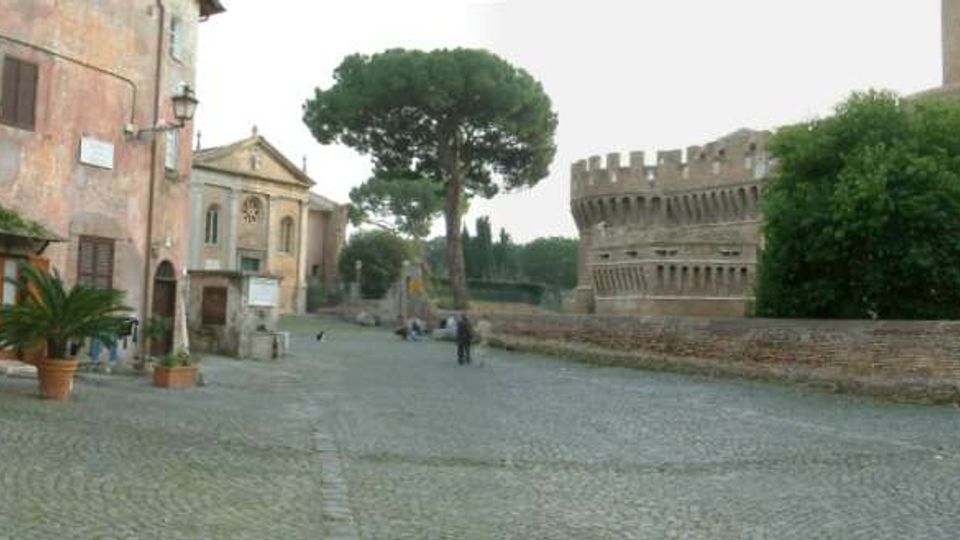Ampia piazza lastricata in pietra accanto a edifici storici, con una facciata intonacata e vasi di piante sul lato sinistro e una piccola chiesa sullo sfondo. A destra si vedono mura in mattoni con torre merlata e una grande chioma di alberi, tra cui un pino marittimo al centro.