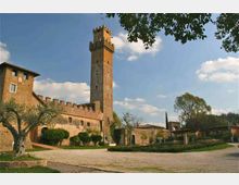 Castello in pietra e mattoni con alta torre merlata, affiancato da mura con camminamenti e archi. Davanti si estende un cortile ghiaioso con giardini curati e alberi, sotto un cielo azzurro con nuvole.