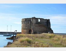 Rovine di una torre o fortificazione in pietra su un promontorio erboso, affacciata su un porto con acqua calma e una diga frangiflutti sullo sfondo. La struttura è cilindrica e parzialmente crollata, con aperture murate e muri irregolari.