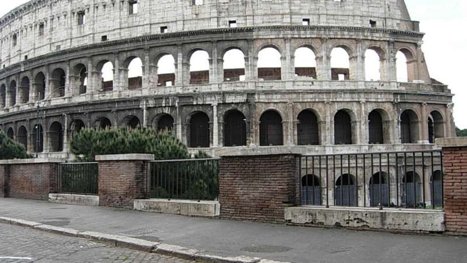 Veduta esterna del Colosseo, anfiteatro romano in pietra con più ordini di archi sovrapposti e aperture. In primo piano si vedono un marciapiede con pavé, una ringhiera metallica e muretti in mattoni con vegetazione bassa.