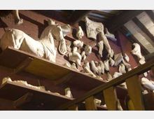 Interno di un laboratorio o magazzino con scaffalature in legno cariche di frammenti di sculture e calchi in gesso, tra cui teste e arti, e una grande testa di cavallo. Sullo sfondo si vede un soffitto con travi e pannelli, illuminato da luce artificiale.