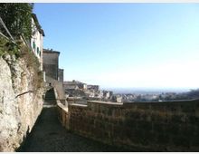 Stradina in salita lastricata lungo una parete rocciosa e case in pietra, affiancata da un muretto di contenimento. Sullo sfondo si vede un borgo collinare e un ampio panorama di campagna fino all’orizzonte sotto un cielo limpido.