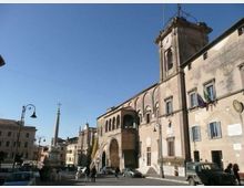 Piazza cittadina con grande edificio storico in pietra e mattoni, con archi, finestre e una torre campanaria laterale, affacciato sulla strada. Sullo sfondo si vede un obelisco con croce e lampioni, con auto parcheggiate e altri palazzi lungo la piazza sotto cielo sereno.