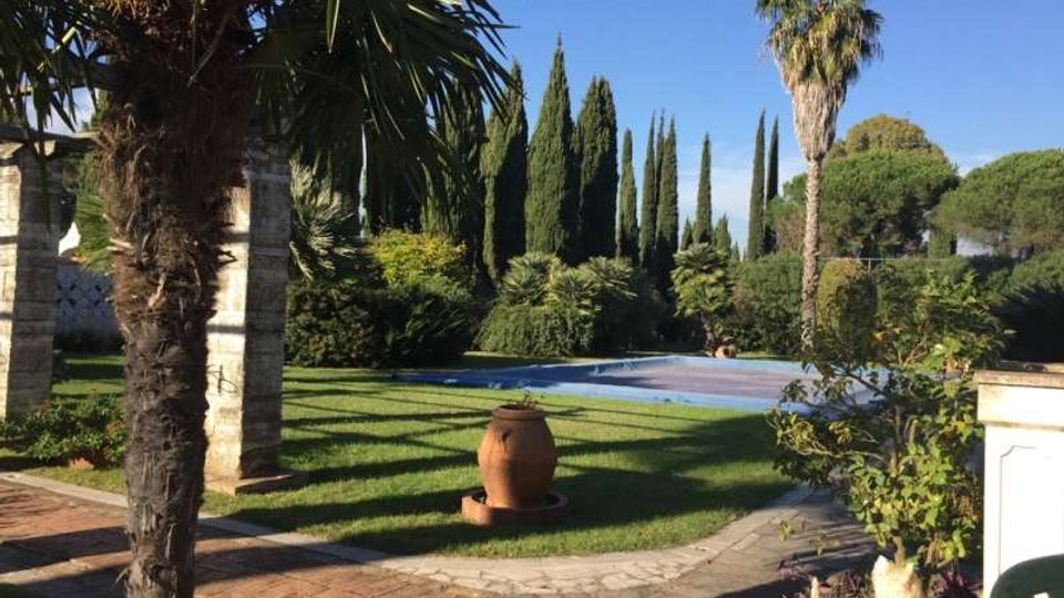 Giardino con prato curato e vialetti in pietra e mattoni, con una piscina rettangolare sullo sfondo. L’area è circondata da palme, cipressi e siepi, con un grande vaso in terracotta al centro e un cielo sereno.