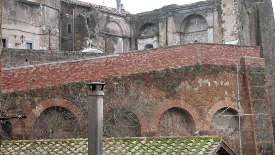 Rovine di un complesso storico in muratura con grandi arcate e nicchie, affiancate da edifici in pietra. In primo piano si vede un muro di contenimento in mattoni sopra una struttura ad archi e tetti con tegole verdi.