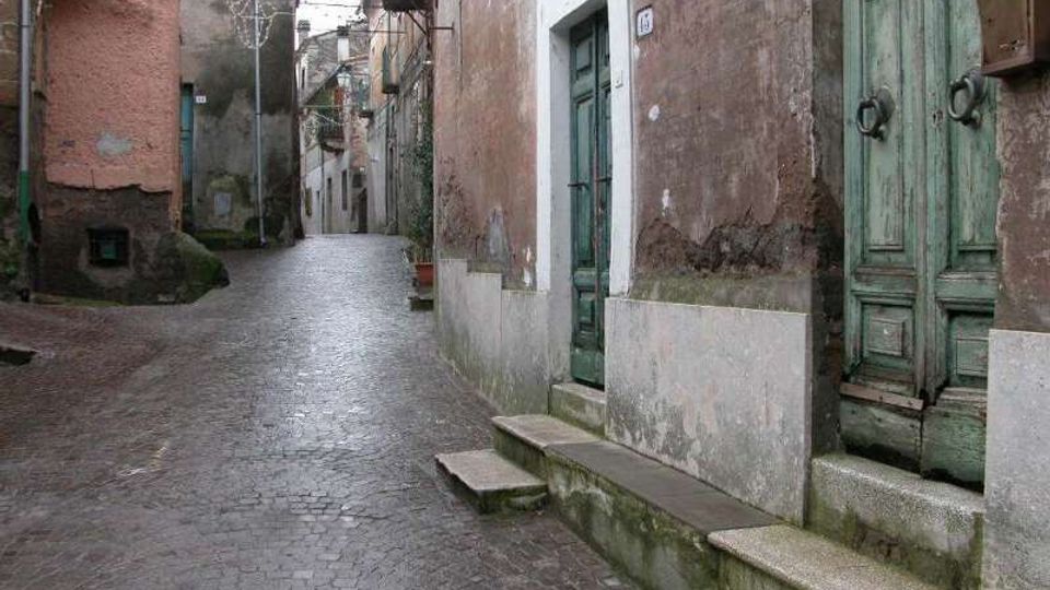 Vicolo stretto con pavimentazione in pietra, fiancheggiato da edifici antichi con intonaco usurato e portoni in legno verde con piccoli gradini d’ingresso. In alto si vedono balconi in ferro e cavi sospesi lungo la strada che curva tra le case.