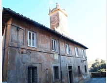 Edificio in muratura intonacata e consumata con tetto a falde e finestre rettangolari, affacciato su un panorama collinare. Dietro la copertura si innalza una torre con quadrante di orologio e piccoli elementi decorativi sulla sommità.