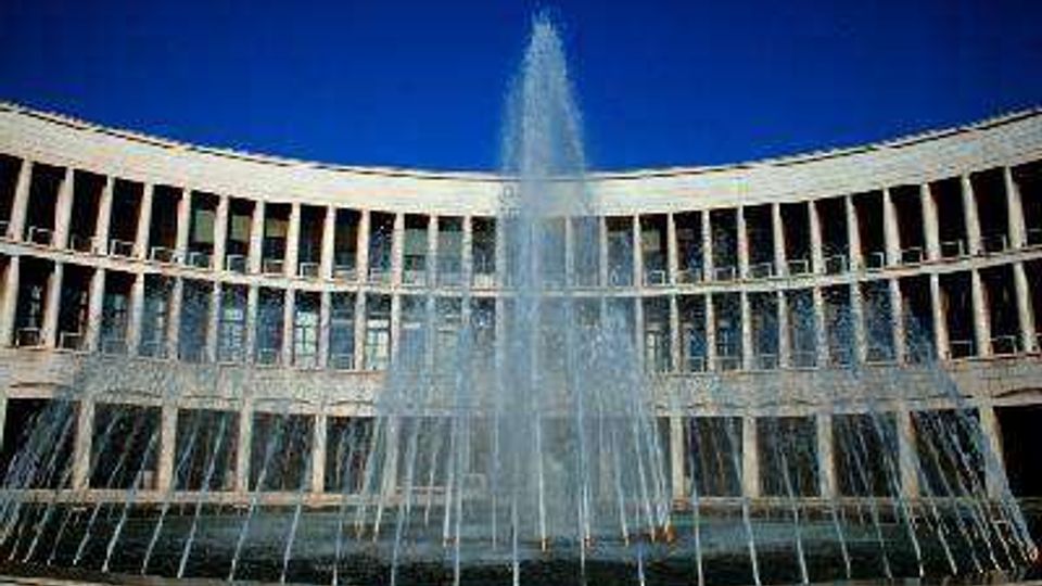Fontana con alto getto d’acqua al centro di una piazza, davanti a un edificio moderno semicircolare con facciata a colonne e grandi finestre. Sullo sfondo si vede il cielo azzurro.