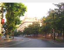 Viale urbano ampio e curvo, fiancheggiato da alberi e lampioni, con una recinzione lungo il marciapiede. Sullo sfondo si vede un grande edificio storico chiaro con facciata decorata e cupola.
