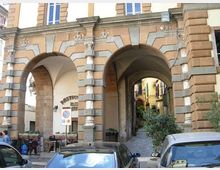Facciata di un edificio in stile storico con grandi arcate in pietra e muratura bugnata, che formano un portico con volte e un passaggio pedonale verso una stretta via interna. Davanti alle arcate si vedono auto parcheggiate e, sotto il portico, arredi e insegne di un locale.
