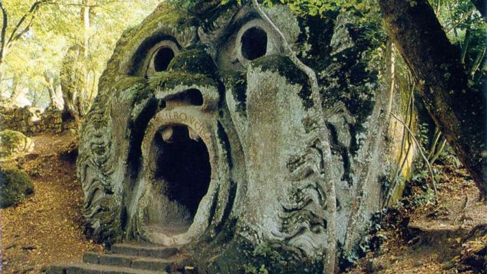 Scultura in pietra a forma di volto con grande apertura centrale simile a una bocca e cavità circolari come occhi, parzialmente coperta di muschio. Si trova in un’area boschiva, con piccoli gradini che conducono all’ingresso e alberi e sentieri tutt’intorno.
