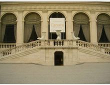 Facciata di un edificio storico con grandi finestre ad arco e grate in ferro, affacciata su una scalinata simmetrica in pietra con balaustra. Al centro si vede un ingresso sottostante e un grande cartellone espositivo con la scritta “Canova”.