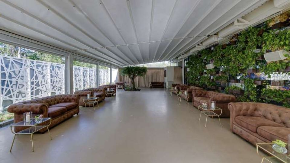 Ampia lounge coperta con soffitto a travi bianche e pavimento chiaro, arredata con divani in pelle marrone e piccoli tavolini. Lungo un lato ci sono grandi vetrate e pannelli schermanti geometrici, mentre sull’altro si sviluppa una parete verde con piante e mensole.
