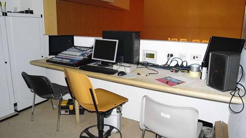 Piccola sala di controllo con banco attrezzato con mixer audio, computer e monitor, cavi e altoparlanti. Sono presenti sedie e armadi laterali, con pareti interne in legno e arancione e un proiettore fissato al soffitto.