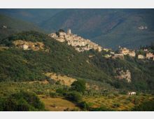 Borgo medievale arroccato su una collina, con case in pietra e una torre campanaria, circondato da pendii boscosi e terrazzamenti agricoli. Sullo sfondo si vedono montagne verdi e una valle ampia.