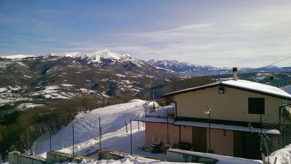 Casa di montagna con tetto innevato e recinzione metallica in primo piano, su un pendio coperto di neve. Sullo sfondo si vede un’ampia catena di montagne con cime innevate sotto un cielo sereno.