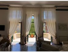 Interno di un salone luminoso con tre alte porte-finestre ad arco, coperte da tende bianche, che si aprono su una terrazza con balaustra in pietra e un grande vaso. Pavimento piastrellato decorato e arredi da soggiorno con divani; all’esterno si vede vegetazione e alcuni edifici vicini.