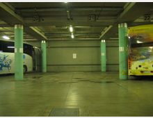 Interno di un’autorimessa o terminal bus sotterraneo con pavimento in cemento lucido, colonne cilindriche verdi e illuminazione a neon sul soffitto. Si vedono due autobus parcheggiati ai lati e numeri di stallo (20, 21, 22) segnati sulle travi superiori.