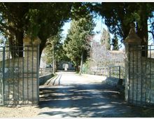 Ingresso con cancello in ferro aperto tra pilastri in mattoni, che immette in un vialetto asfaltato. Il percorso è fiancheggiato da ringhiere e alti alberi sempreverdi, con edifici sullo sfondo.