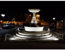 Fontana in pietra illuminata di notte, con vasca circolare su gradoni concentrici e un’alta coppa centrale. Intorno si vede una piccola piazza/rotonda con catene di protezione, auto parcheggiate e lampioni lungo la strada in salita.