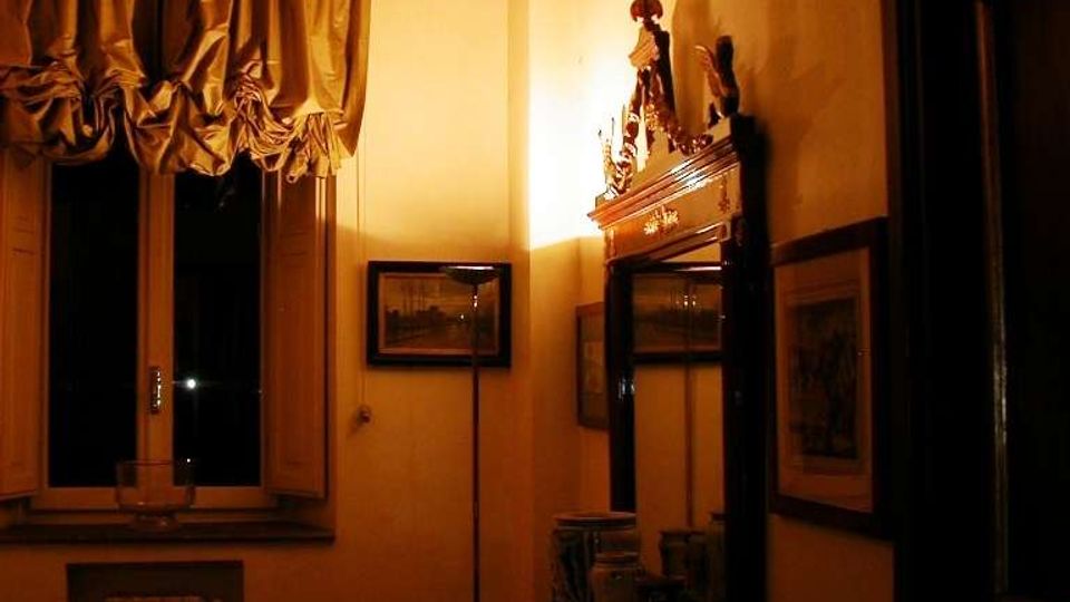 Interno di una stanza in stile tradizionale con soffitto a travi in legno, finestra con tende arricciate e pareti illuminate da luce calda. In primo piano un tavolo con oggetti decorativi e uno specchio incorniciato sopra, con quadri appesi e una porta scura sul lato destro.