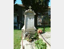 Monumento funerario in marmo in un cimitero, con croce in cima e lapide incisa su un basamento rialzato. Intorno si vedono prato, fiori e altre tombe con loculi sullo sfondo, parzialmente ombreggiate da alberi alti.