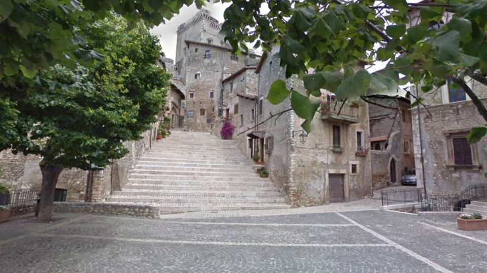 Scalinata in pietra che sale da una piazza lastricata verso un borgo medievale con edifici in muratura e una torre centrale. Alberi e rami in primo piano incorniciano la scena, con case addossate ai lati della scalinata.