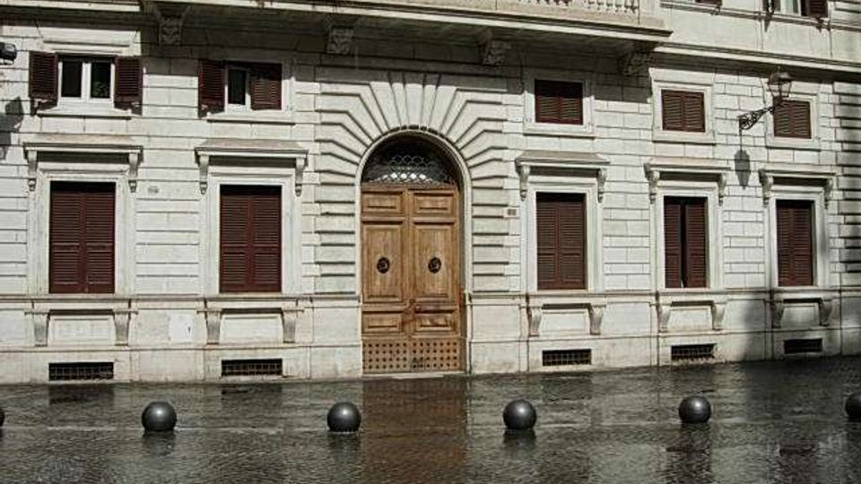 Facciata di un edificio storico in pietra chiara con portale centrale ad arco in legno, finestre con persiane scure e balcone con balaustra. In primo piano una piazza pavimentata con dissuasori sferici neri e superficie bagnata che riflette l’edificio.