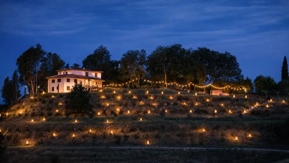 Casa di campagna su una collina al crepuscolo, circondata da alberi e da una fila di luci sospese. Il pendio è punteggiato da numerose lanterne accese disposte lungo i terrazzamenti.