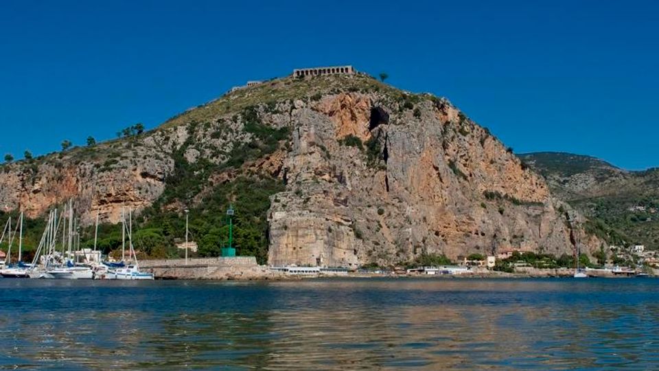 Veduta di una baia con acqua calma e riflessi in primo piano, con un promontorio roccioso e scogliere sullo sfondo. Alla base si vede un piccolo porto con barche a vela e un faro verde su un molo.