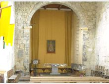 Interno di una piccola chiesa in pietra con grande arco centrale e pareti in muratura a vista. In fondo si vedono l’altare semplice, una pedana e sedie in legno disposte lungo i lati, con un dipinto appeso sulla parete posteriore.