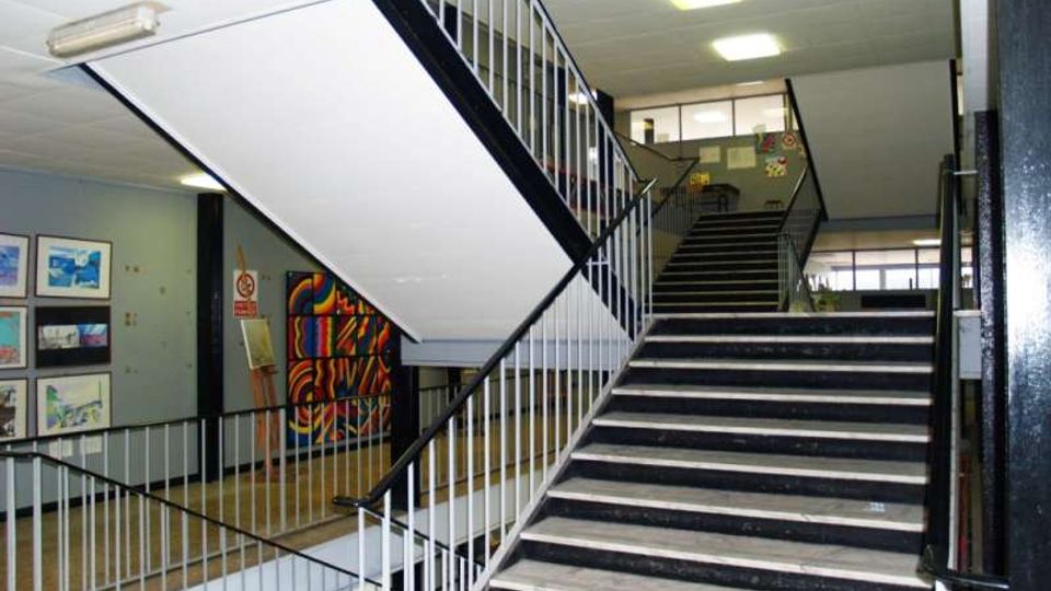 Atrio interno di un edificio con ampia scala in cemento e ringhiere metalliche bianche e nere, illuminato da plafoniere a soffitto. Sulle pareti del piano inferiore si vedono quadri e un grande murale colorato, con finestre sul fondo.