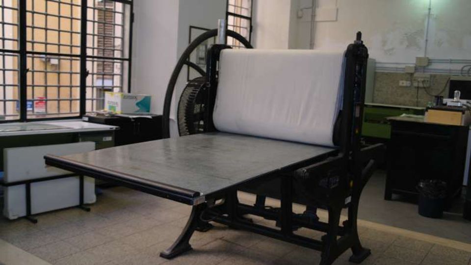 Interno di un laboratorio con grandi finestre a griglia e pavimento piastrellato, arredato con tavoli e attrezzature. In primo piano si vede una grande macchina da stampa con rullo e piano di lavoro metallico.