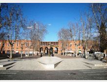Piazza urbana pavimentata con gradoni e una piattaforma centrale rialzata, circondata da alberi spogli. Sullo sfondo si vede un grande edificio in mattoni rossi con una facciata simmetrica e un porticato ad archi.