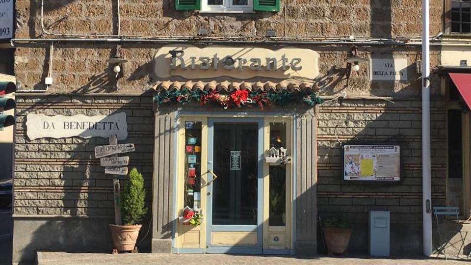 Facciata in pietra di un ristorante con ingresso centrale a porta vetrata, insegna “Ristorante” e decorazioni floreali sopra la vetrina. Ai lati ci sono vasi con piante, cartelli e una bacheca informativa, con finestre superiori dotate di persiane verdi.