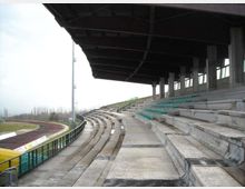 Tribuna coperta di uno stadio con gradinate in cemento e file di sedili verdi, affacciata su una pista di atletica curva. Struttura con tettoia scura sostenuta da pilastri e parapetti metallici lungo il bordo della pista.