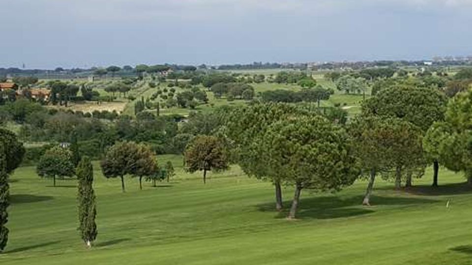 Ampio campo da golf con prati verdi ondulati e alberi sparsi, con alcuni cipressi in primo piano. Sullo sfondo si vede una campagna pianeggiante con edifici lontani sotto un cielo parzialmente nuvoloso.
