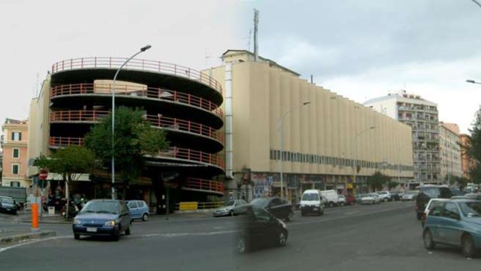 Ampio incrocio urbano con una lunga struttura beige a più piani e, sul lato sinistro, un parcheggio multipiano circolare con rampe esterne e ringhiere rosse. Lungo la strada si vedono auto, semafori e palazzi residenziali sullo sfondo.