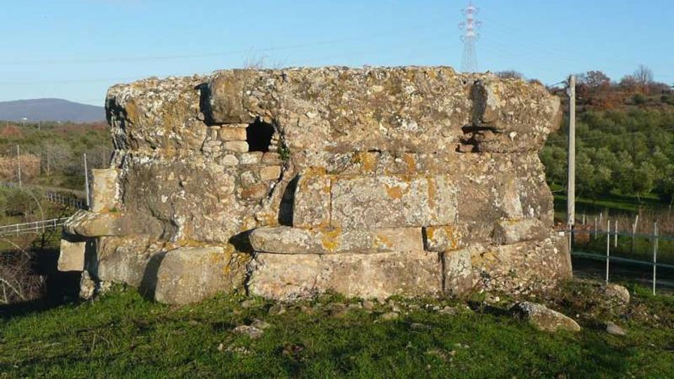 Rovine di una piccola costruzione in pietra con muri spessi e aperture irregolari, situata su un prato. Sullo sfondo si vedono campi e alberi su colline, con un traliccio e pali elettrici.