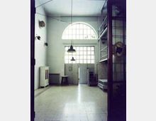 Interno di un ampio spazio industriale con pavimento piastrellato, pareti chiare e grandi finestre a griglia con arco superiore sopra una porta centrale. Lampade a sospensione dal soffitto, scaffalature e una struttura a soppalco in metallo sul lato destro, con radiatore e armadietto sul lato sinistro.