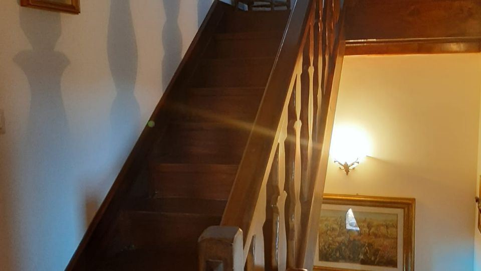Scala interna in legno con corrimano e balaustra intagliata che sale a un pianerottolo superiore. Pareti bianche decorate con quadri e piatti, illuminate da una lampada a parete.