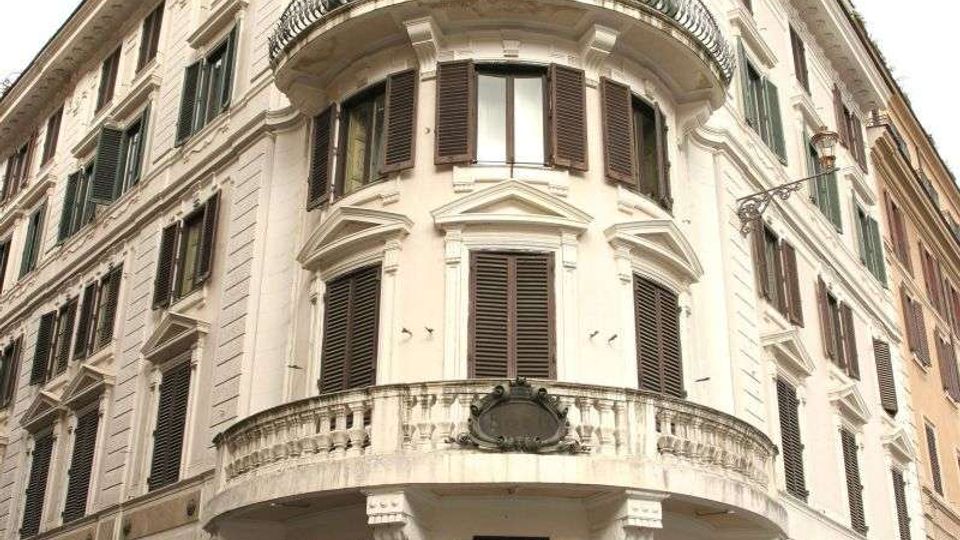 Edificio storico d’angolo in stile neoclassico, con facciata chiara, finestre con persiane scure e balconi curvi con ringhiere in ferro battuto. Al piano terra si trovano vetrine di negozi e ingressi su una strada urbana stretta.