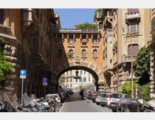 Strada urbana fiancheggiata da alti palazzi storici con facciate in pietra, balconi e finestre ad arco, con auto e moto parcheggiate lungo i lati. In fondo si vede un grande arco in muratura che collega gli edifici e incornicia la vista di un altro tratto di città oltre il passaggio.