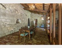 Sala interna in stile rustico con pareti in pietra a vista, soffitto con travi in legno e pavimento in mattoni. Al centro c’è un tavolo da pranzo con sedie, mentre lungo un lato si vede una grande libreria in legno e sul fondo archi e una finestra che illuminano il corridoio.