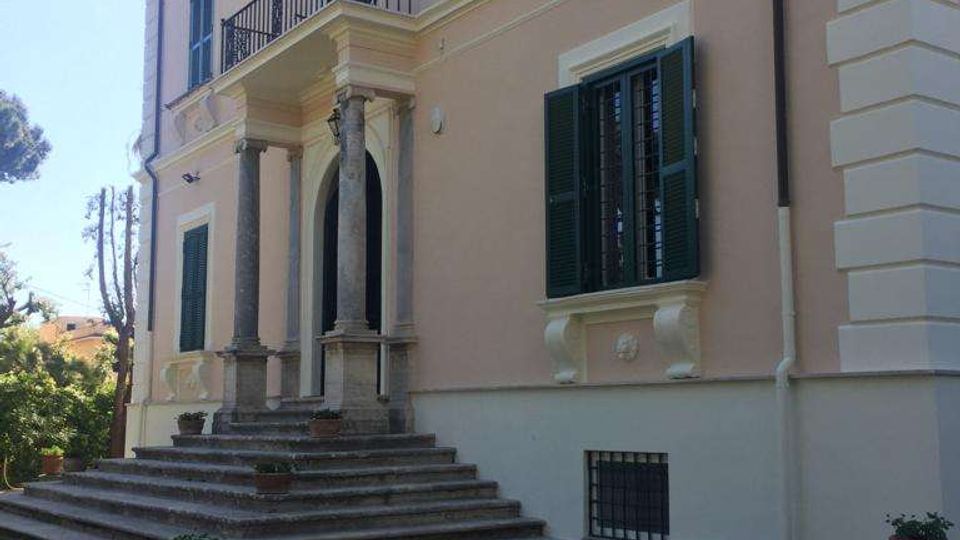 Facciata di una villa storica a due piani con intonaco chiaro, persiane verdi e cornicioni decorativi. Un ampio scalone conduce a un ingresso con portico sorretto da colonne e balconcino, affacciato su un prato con vialetto in pietra.