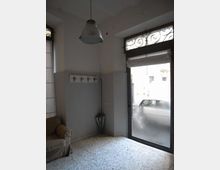 Interno di un piccolo locale con pavimento in graniglia, pareti bianche e una grande porta/vetrina d’ingresso con sopraluce decorato in ferro battuto. Sono visibili una lampada a sospensione, un pannello appendiabiti a parete e una finestra laterale.